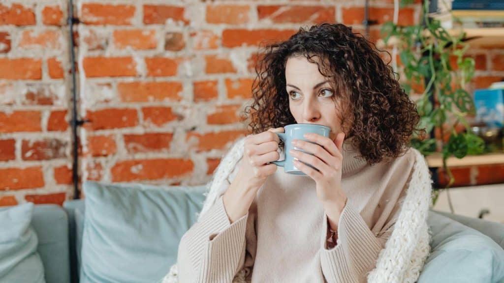 A woman with curly hair drinking from a mug while sitting on a couch.
