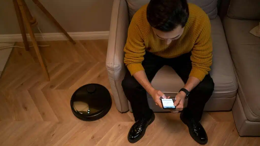 A man sitting on a couch using a smartphone while a robot vacuum rests on the floor nearby.