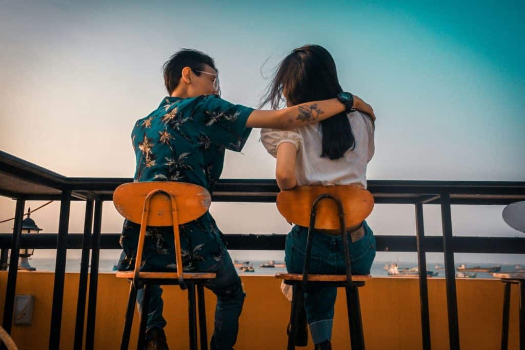 A man and woman sitting on chairs on a balcony overlooking the sea where boats are floating.
