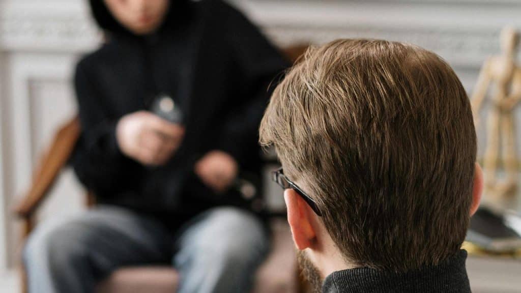 A man in glasses faces another person sitting with a phone.