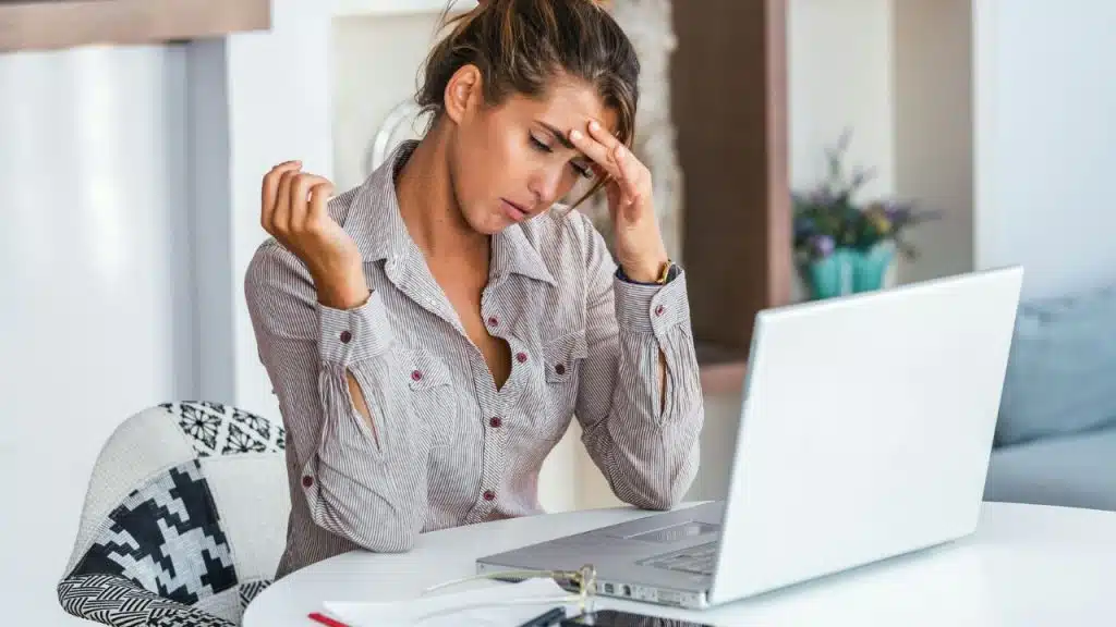 A woman sits at a white table with a laptop, resting her forehead on one hand.