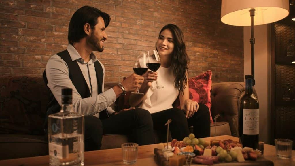 A man and woman clink wine glasses while sitting on a sofa near a table.