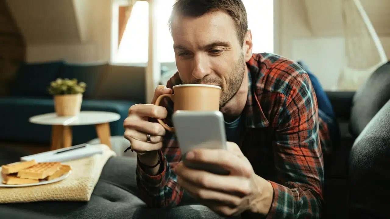 A man lying on a couch drinking coffee and looking at his phone.
