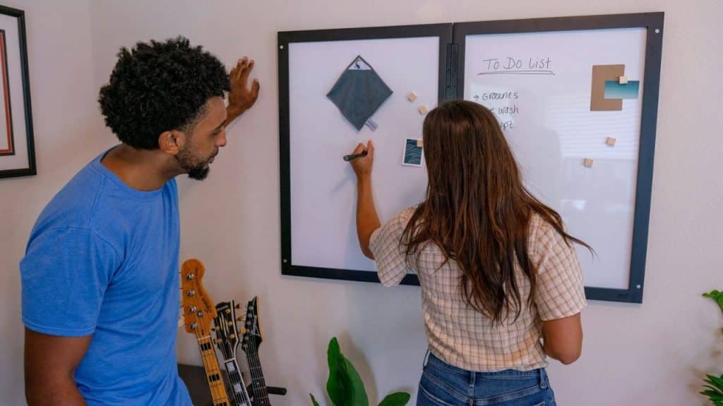 Woman writing on a whiteboard with a man watching, next to a guitar stand.