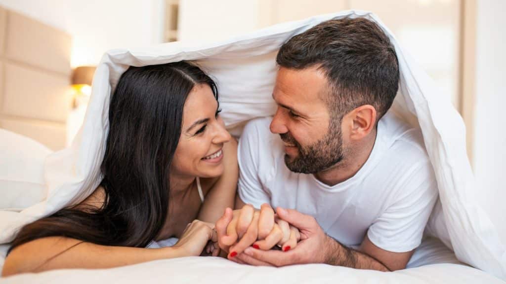 A smiling couple going to kiss under sleeping sheets in the morning.