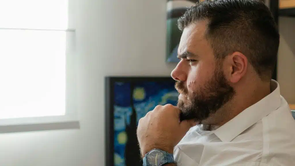 A bearded man in a white shirt and watch is pictured in profile, resting his chin on his hand.