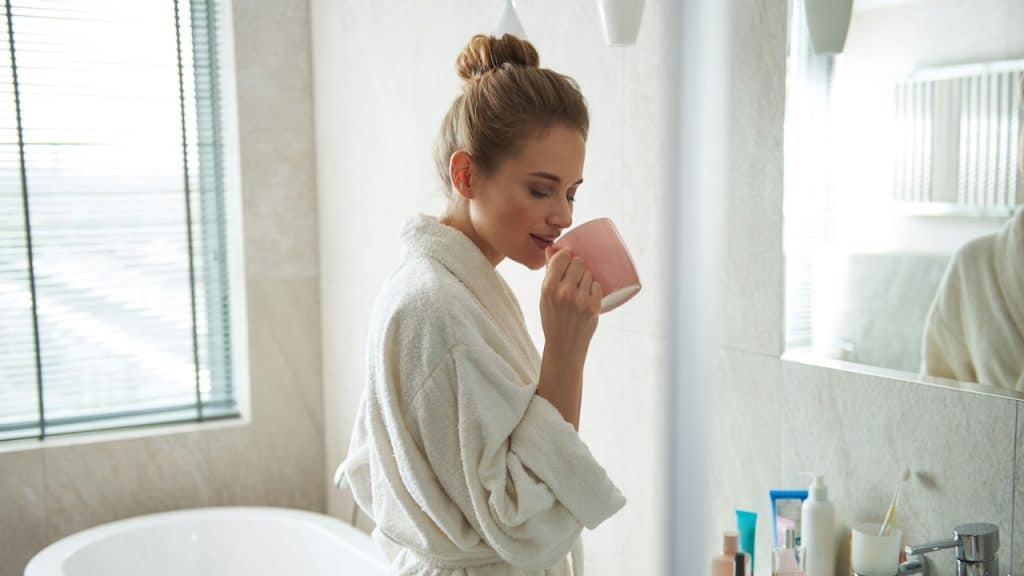 A woman in a white bathrobe stands in a well-lit bathroom, drinking from a pink mug with her hair up in a bun.