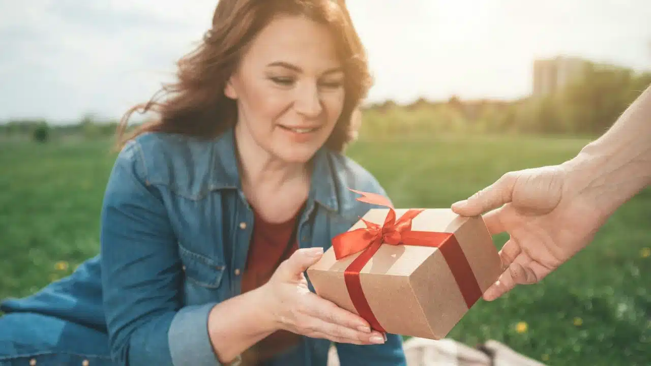 Woman in a denim shirt receiving a gift box tied with a red ribbon in a sunny park.