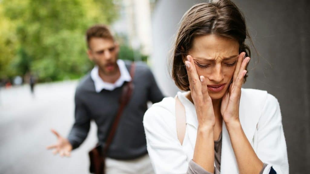 A distraught woman holds her face while a blurred man behind her gestures aggressively outdoors.