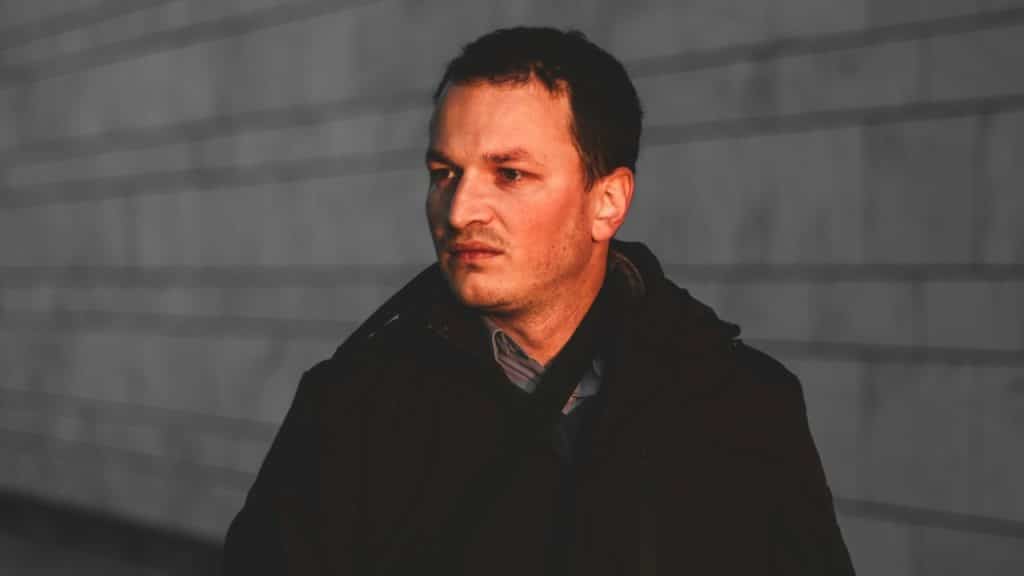 Man in a dark jacket looks off-camera with serious expression against a striped background.