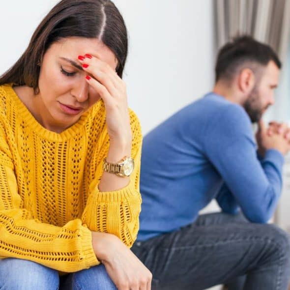 A distressed woman sits on a sofa, holding her forehead, with a man turned away in the background.