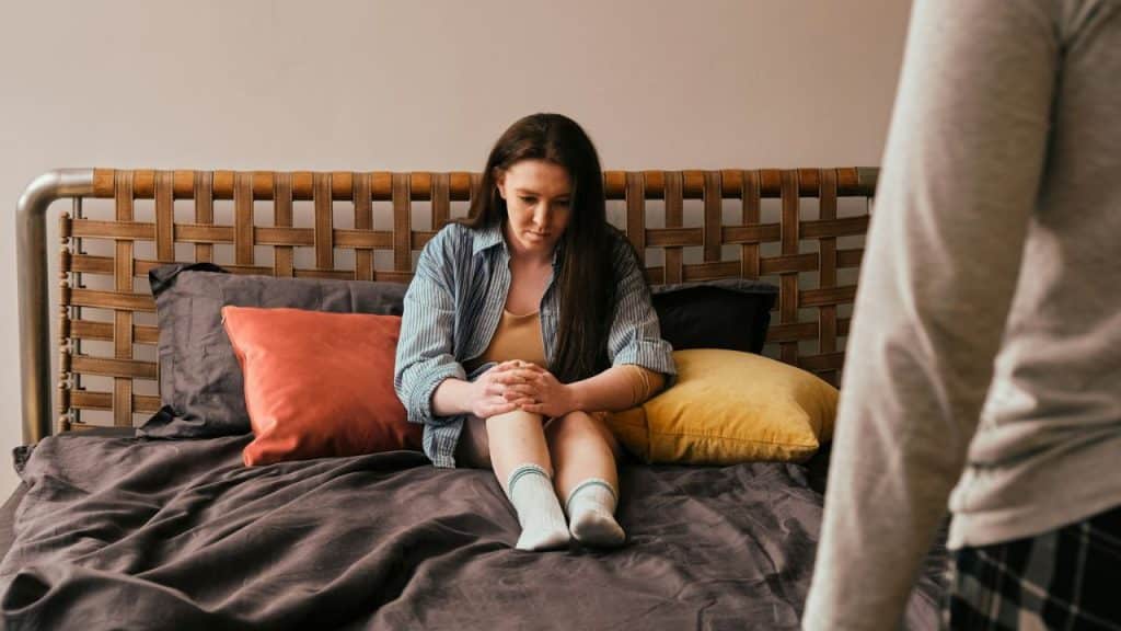 A sad woman sits on a bed with her knees pulled up as a person stands in the foreground.