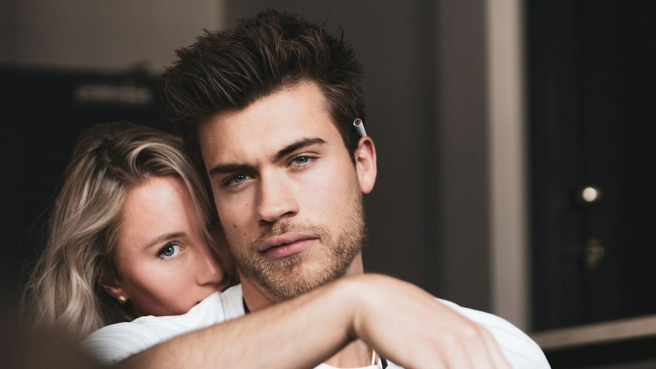 A lightly bearded man looking bored while a woman rests her head on hils shoulder from behind.