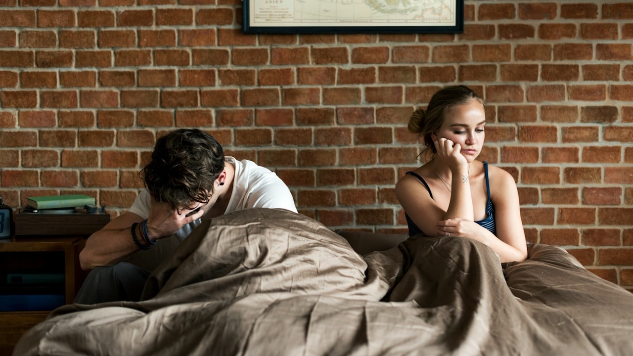 A couple sitting apart in bed, looking upset and avoiding eye contact after a conflict.