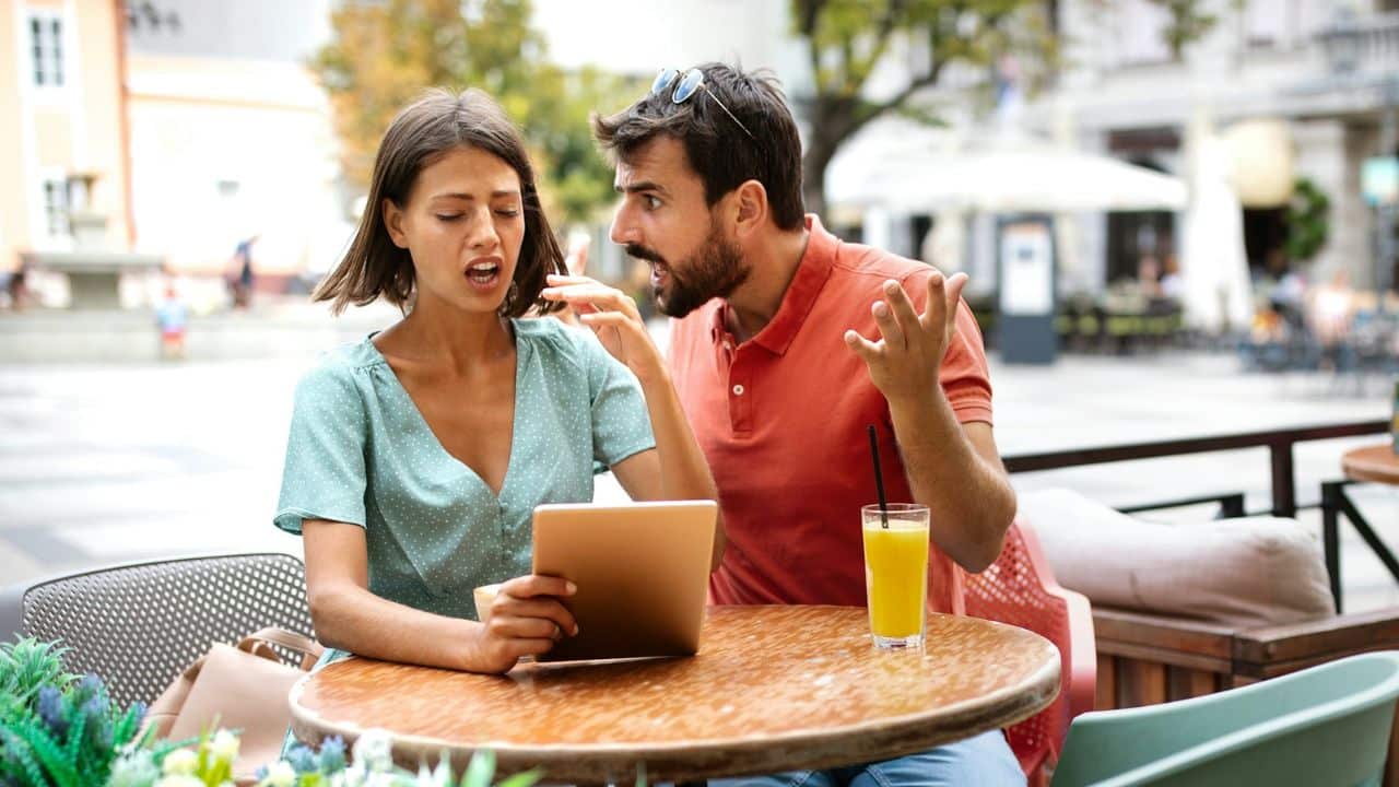 A man shouts with exaggerated gestures at a woman who looks away from a tablet.