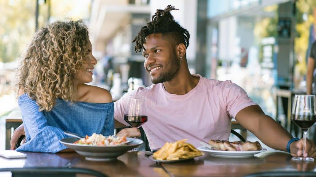 A couple smiles at each other while sharing wine and food at an outdoor restaurant.