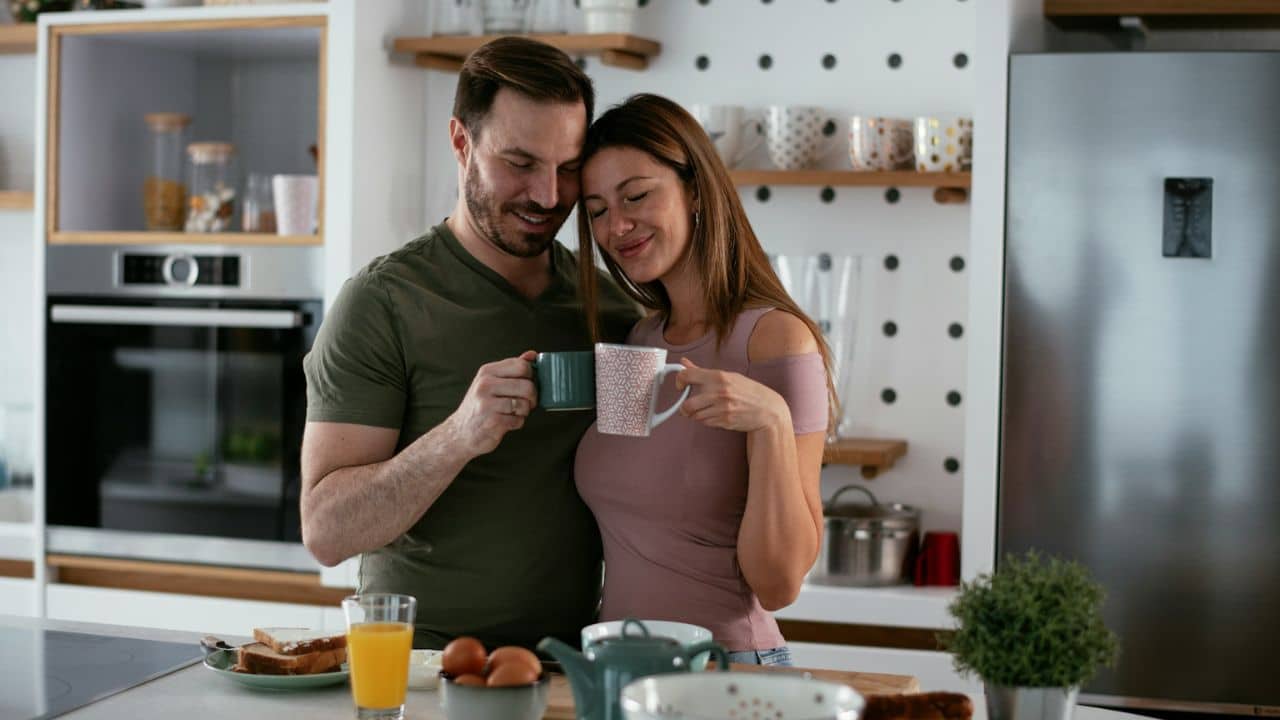 Couple standing close in a kitchen, holding mugs and smiling tenderly.