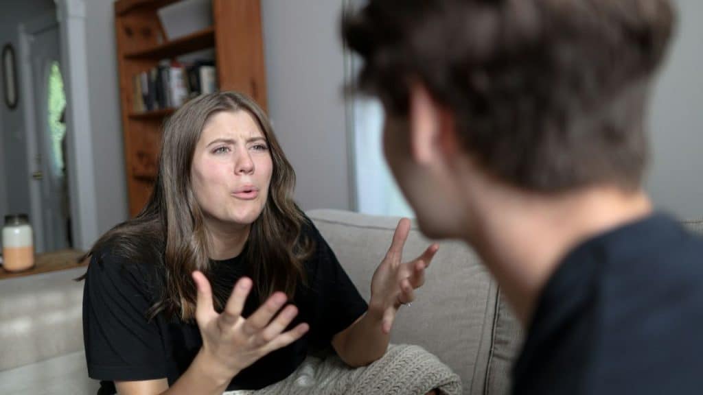 Young woman sitting on a couch gesturing aggressively while talking to a blurred person.