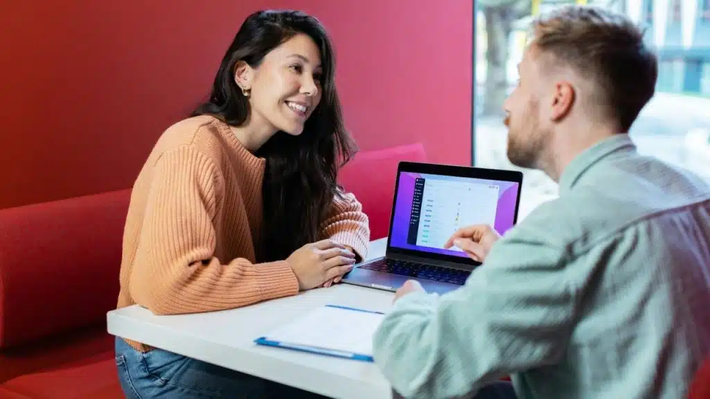 A smiling woman and a man sit at a table, looking at a laptop screen together.