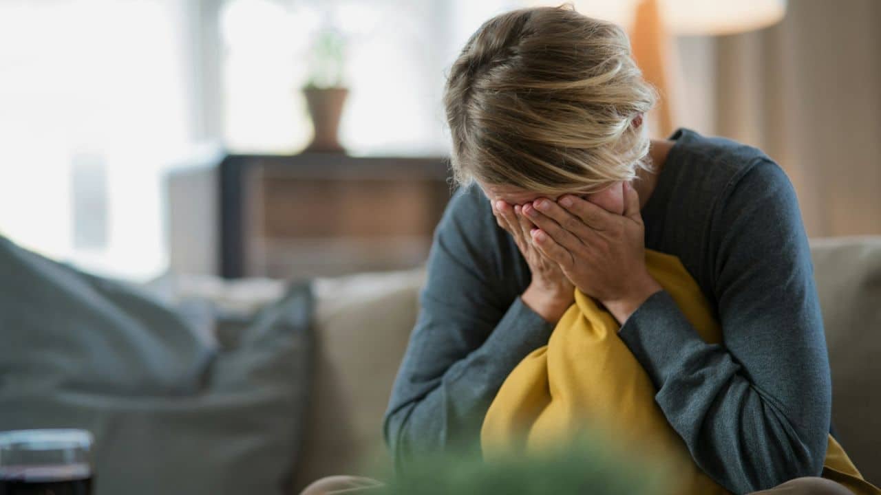 A person sitting on a couch, covering their face with their hands, holding a pillow.