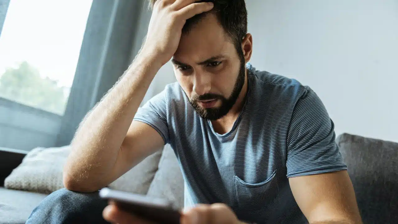 A distressed man with a beard sits on a couch looking intently at his phone.