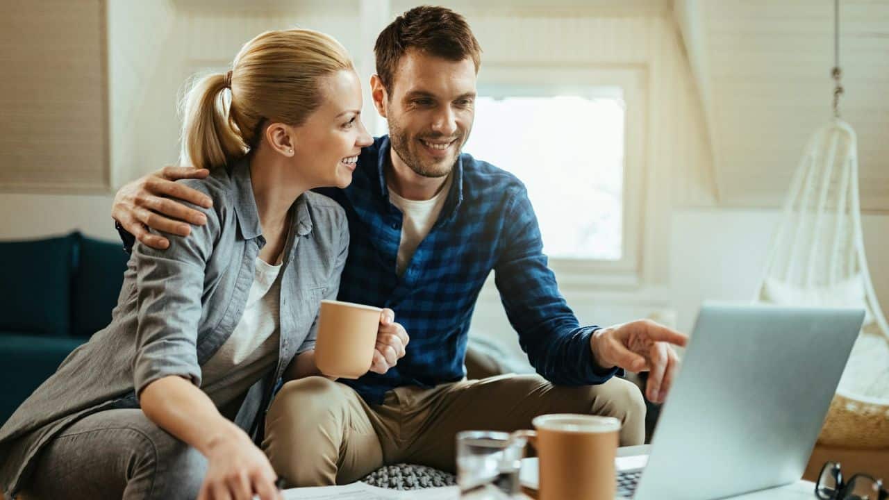 Smiling couple sitting on a couch, looking at a laptop screen together; man points at the screen.