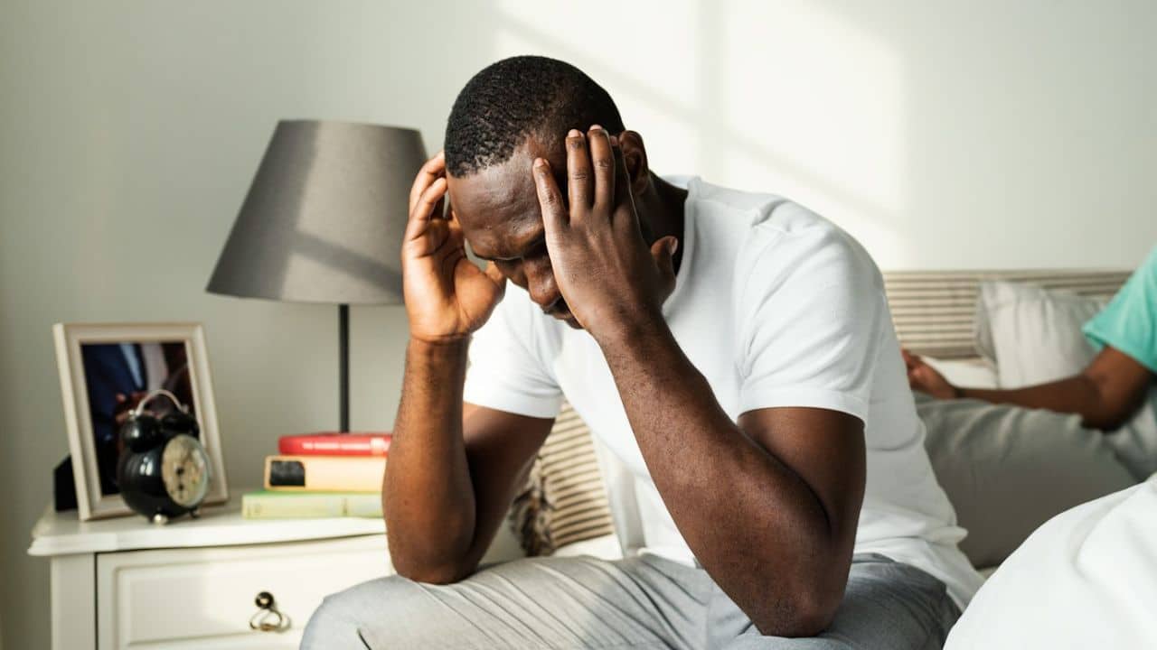 Black man sitting on bed with head in hands, looking down in distress.