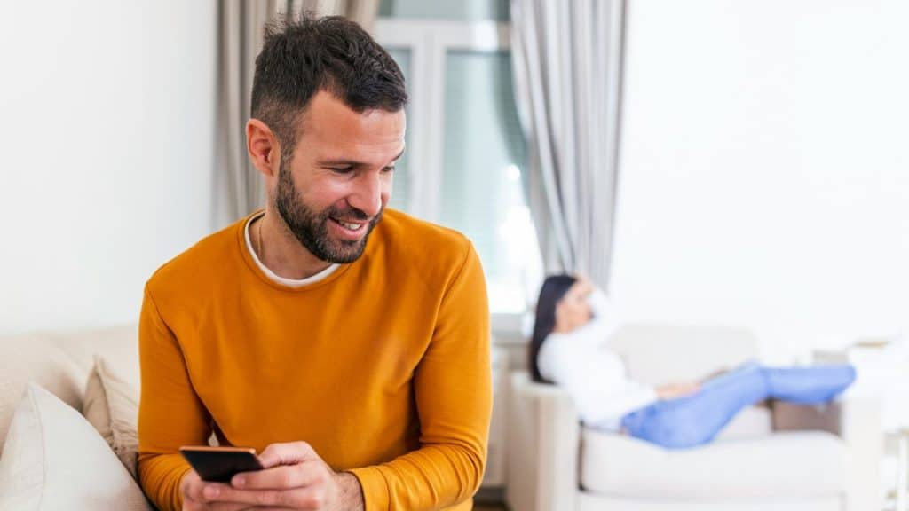 A smiling man uses a phone while a woman relaxes on a sofa in the background.