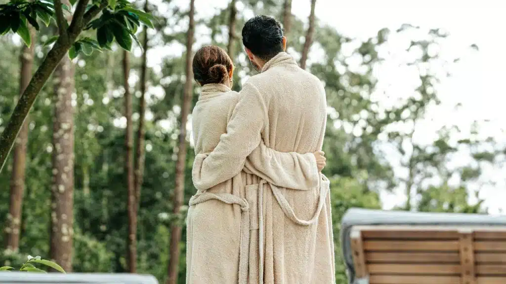 A couple in matching robes embracing while looking at trees outdoors.