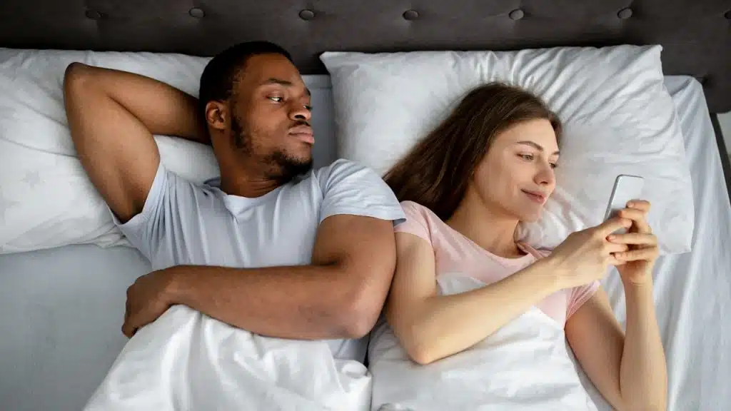 A man staring at a woman’s phone while they lie in a bed together.