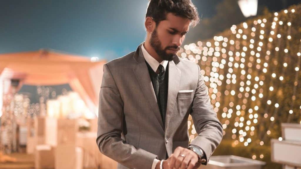 A man in a gray suit looks down at his wristwatch at an outdoor event.