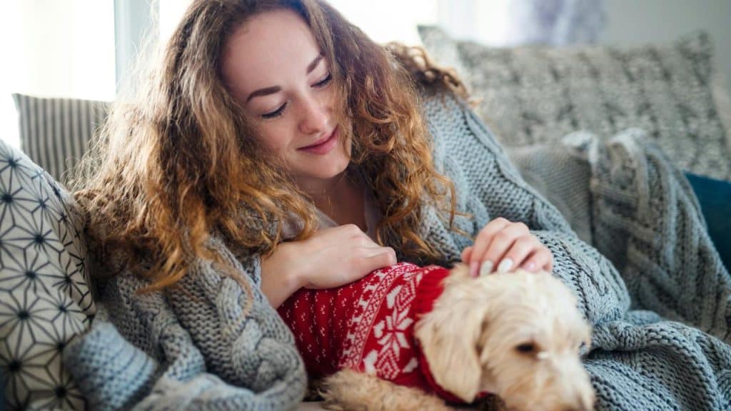 A woman relaxing on a couch while petting a small dog in a sweater.