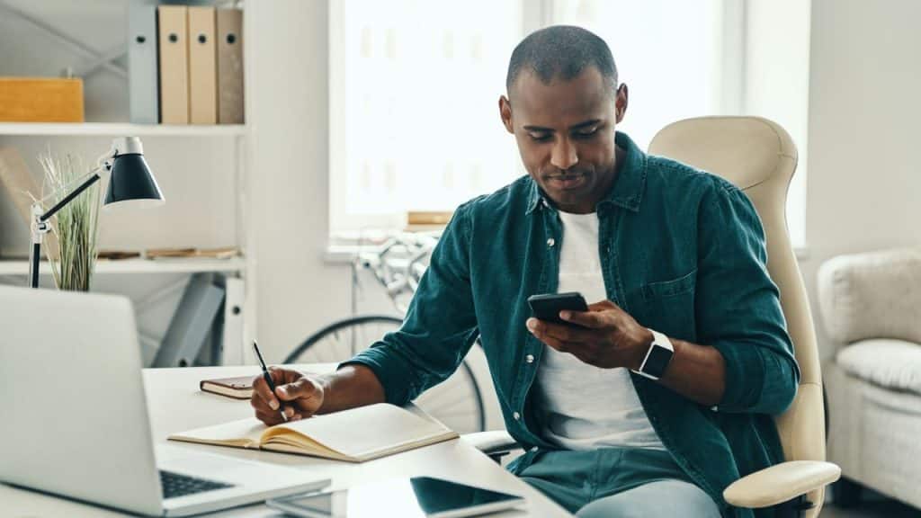 A man seated at a desk holds his phone in one hand and writes in a notebook with the other.
