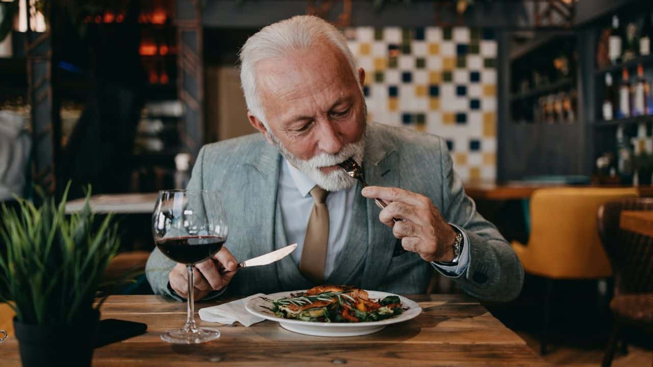 A distinguished older man in a suit eats a plated meal and drinks red wine in a restaurant.