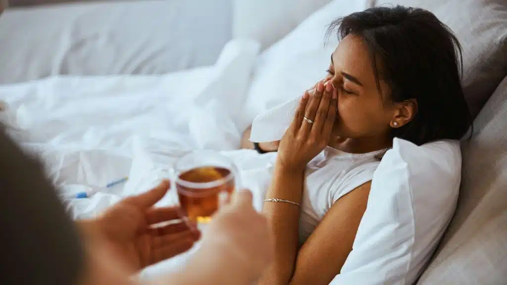 A woman blowing her nose in bed as someone offers her a glass of tea.