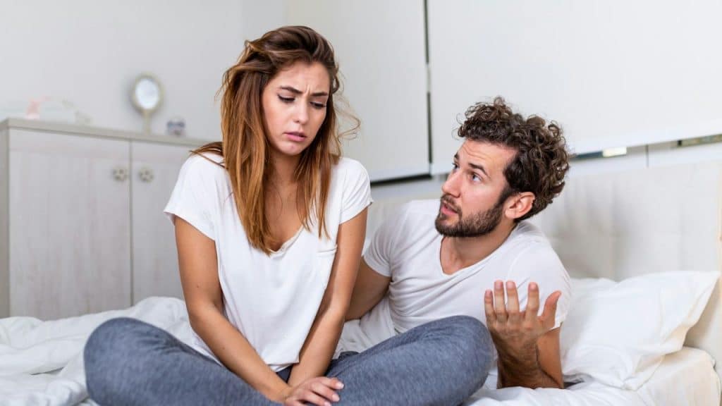 Woman sitting on a bed looking upset while a man gestures and talks to her.