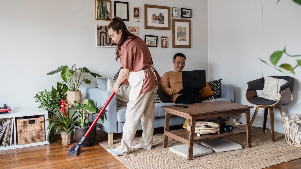 A woman vacuums the floor in a living room while a man sits on the couch using a laptop.