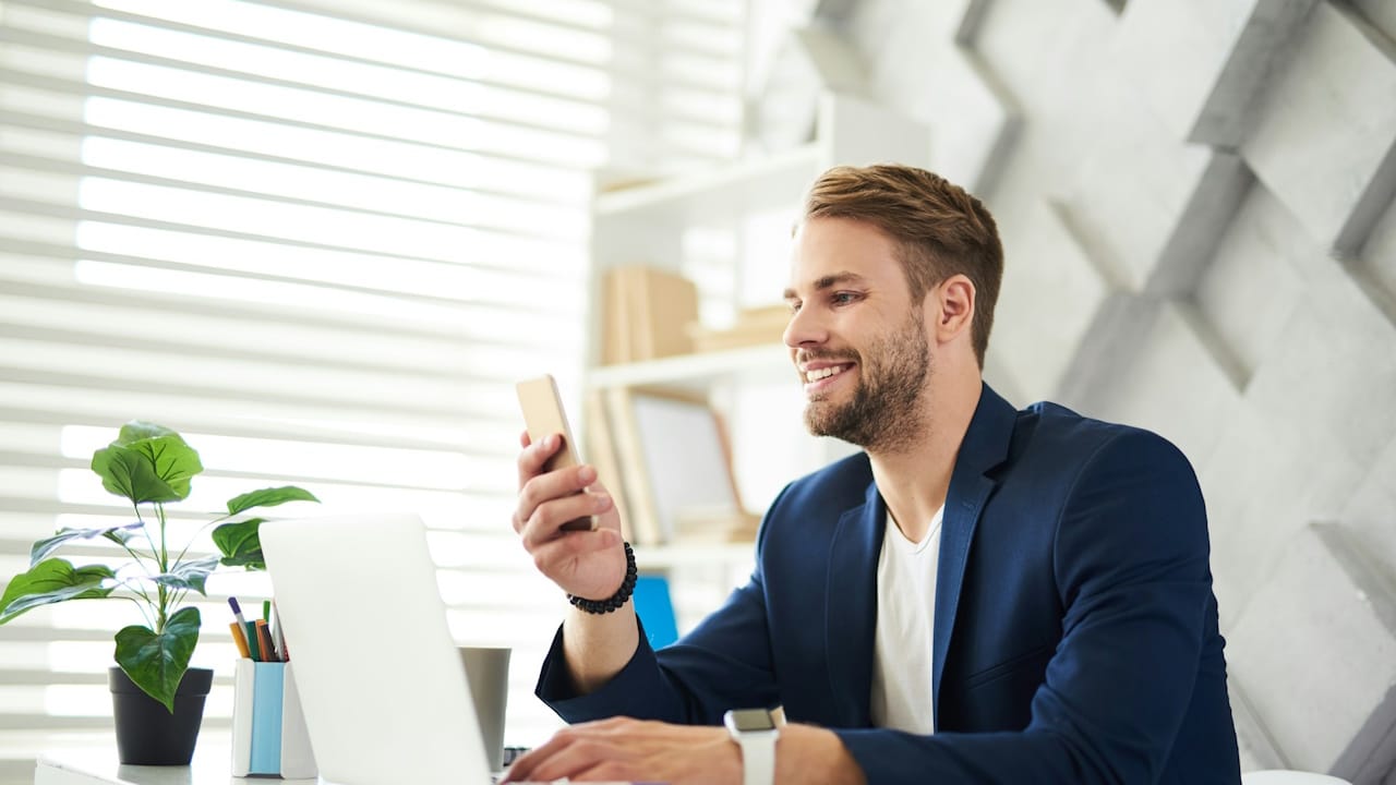 A side view profile of a smiling businessman chatting on the phone in the office; he is sitting at a laptop in front of him and typing on-screen with joy.