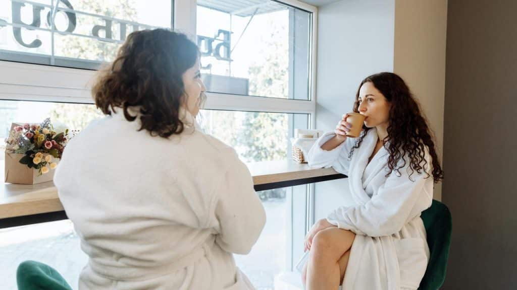 A woman in a robe sipping coffee while talking with another woman by a window.