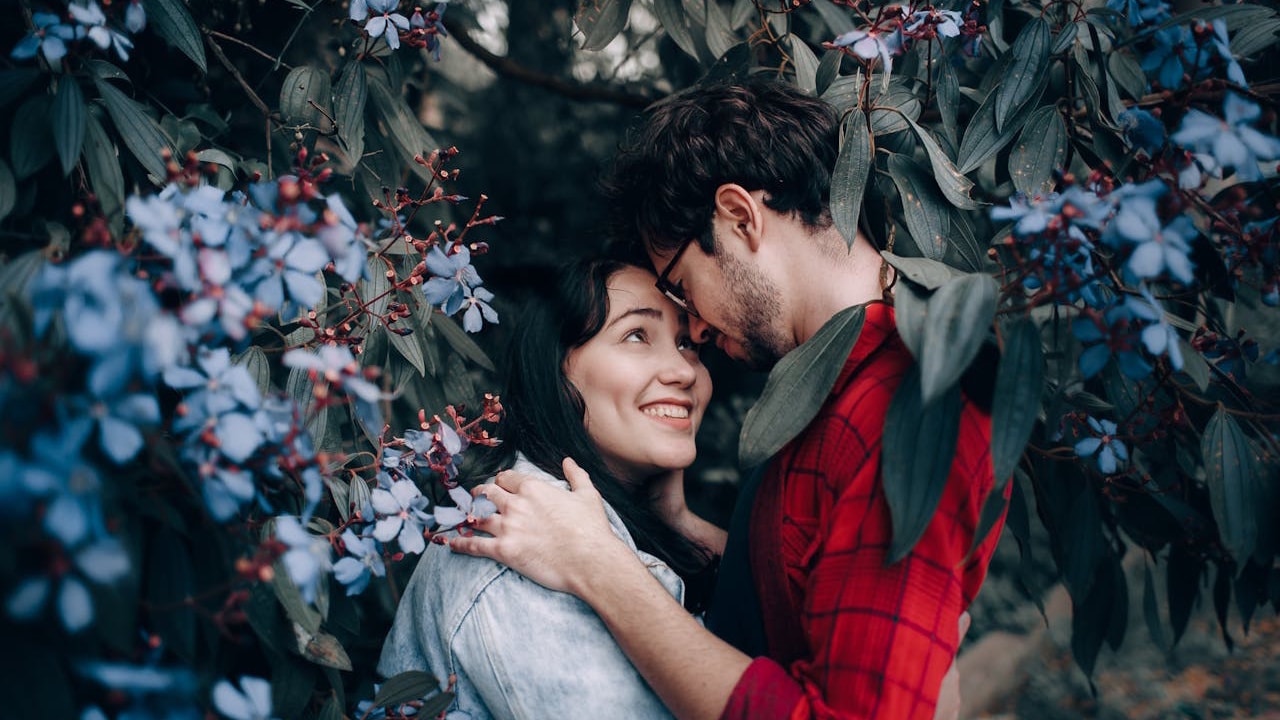 A man and a woman are embracing amidst dark foliage and blue flowers, smiling at each other.
