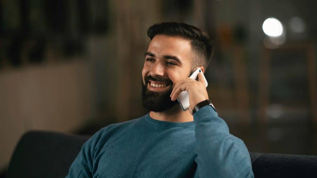 A smiling person sitting on a couch while talking on a smartphone.