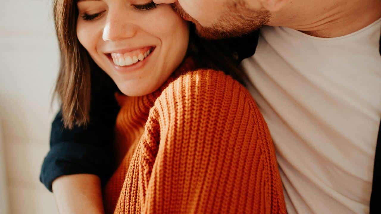 A close-up of a smiling woman being affectionately embraced and kissed on the cheek.