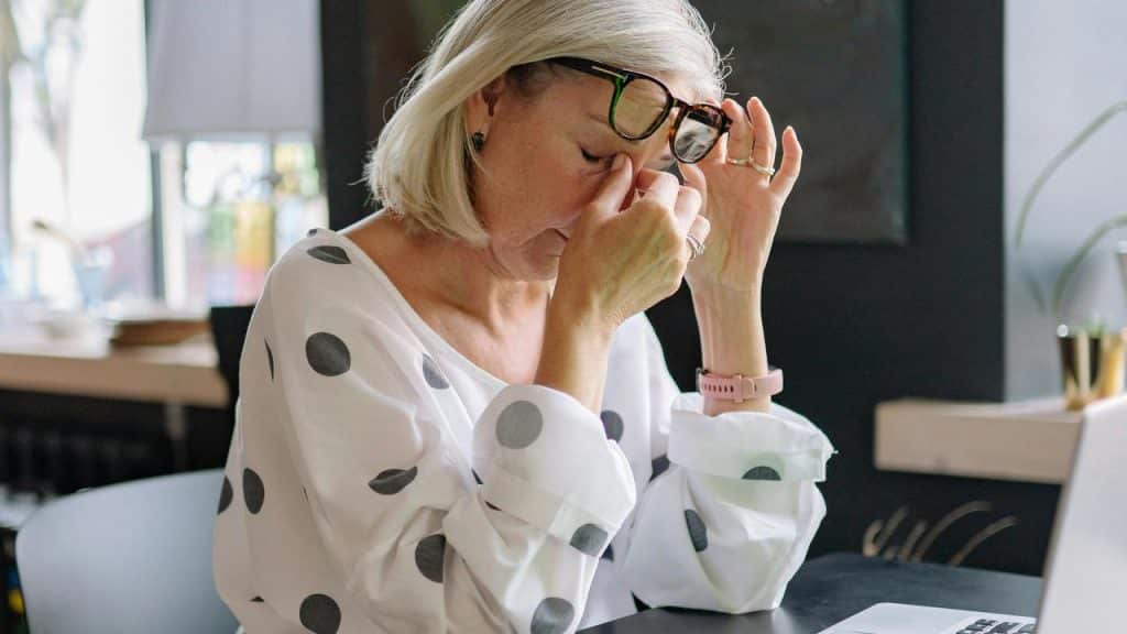 An older woman rubbing her eyes in tiredness at a desk.