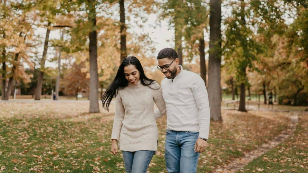 Couple walking through a park in autumn, holding hands and smiling at each other.