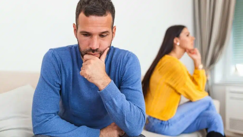 Man in blue looking down thoughtfully, woman in yellow and blue sitting with her back to him.