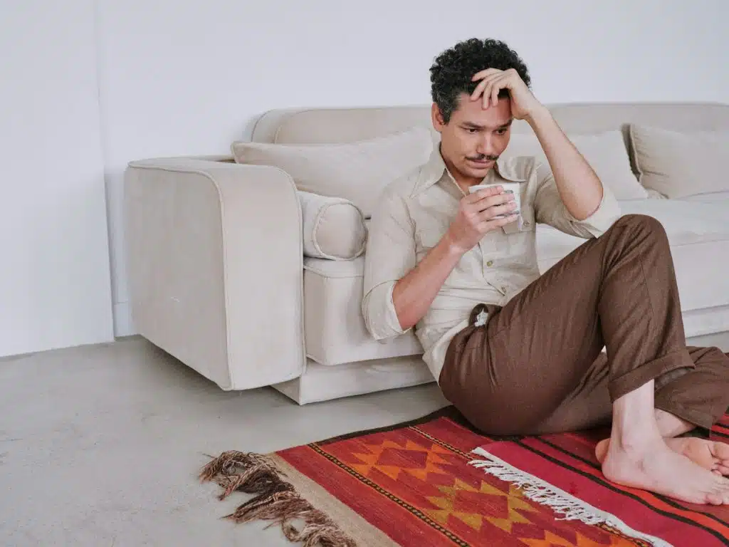 A sad man sitting on the floor while holding a cup of coffee.