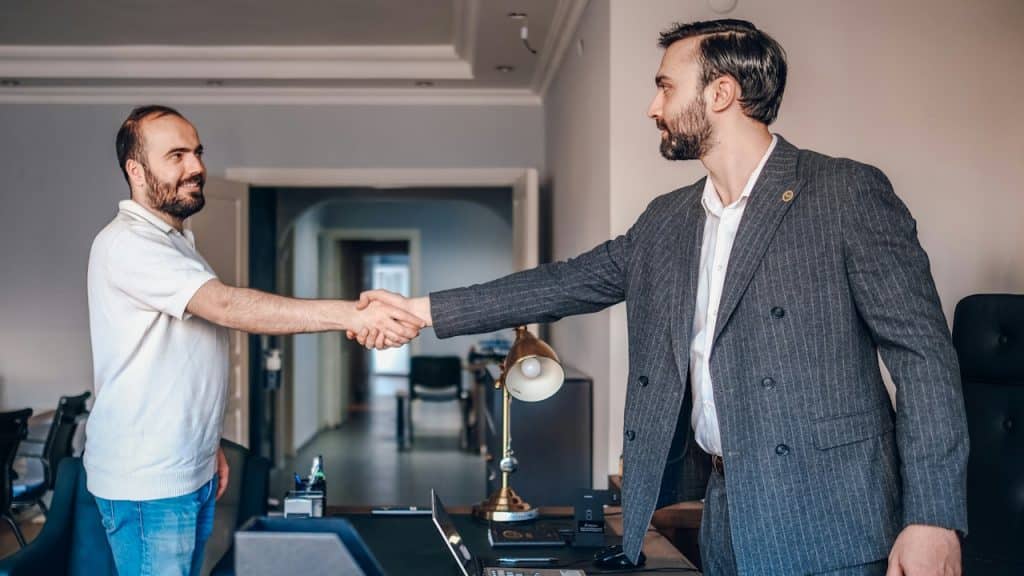 Two men shaking hands across a desk in an office.
