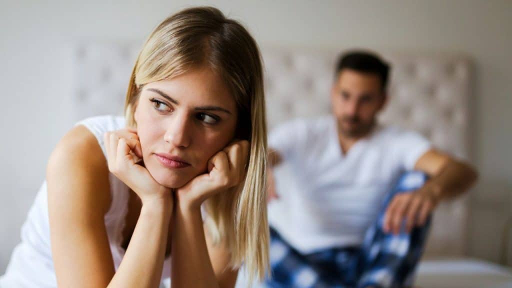 A thoughtful woman sits on a bed with a blurred man in the background.