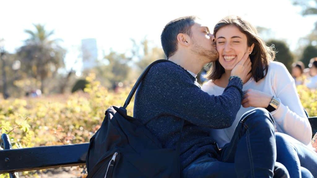 A man kisses a woman on the cheek as they sit together outdoors and smile.