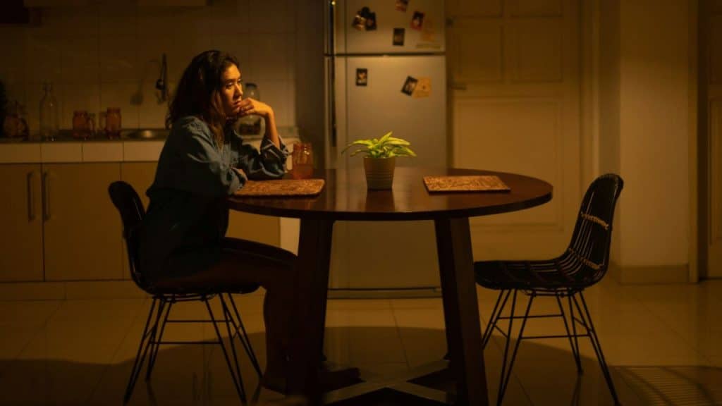 A woman with her chin resting on her hand sits alone at a dark kitchen table.